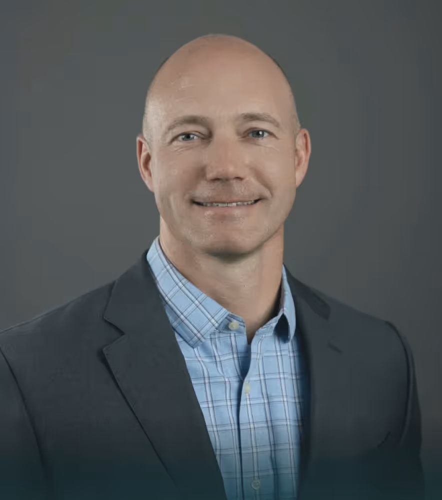 Smiling bald man wearing a dark blazer and blue checked shirt against a gray background.
