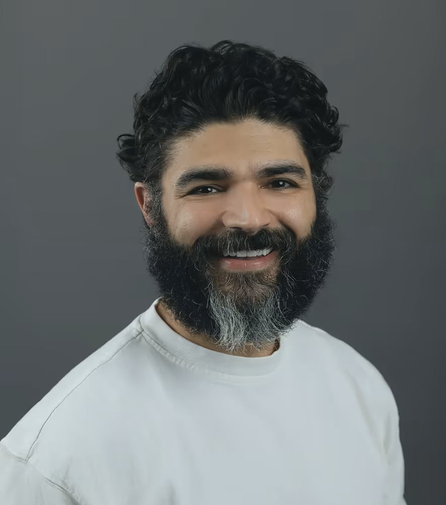 Smiling man with curly dark hair and a full black and gray beard wearing a white shirt against a gray background.