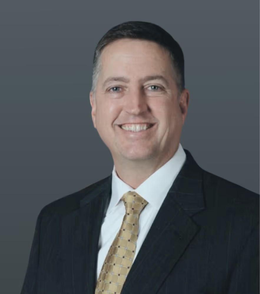 Smiling man in a dark suit, white shirt, and gold patterned tie against a gray background.