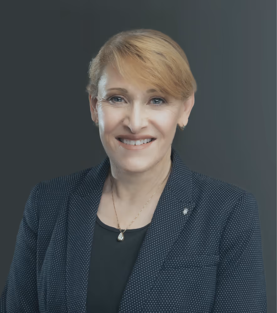Smiling woman with short blonde hair wearing a black polka-dot blazer and a necklace with a teardrop pendant.