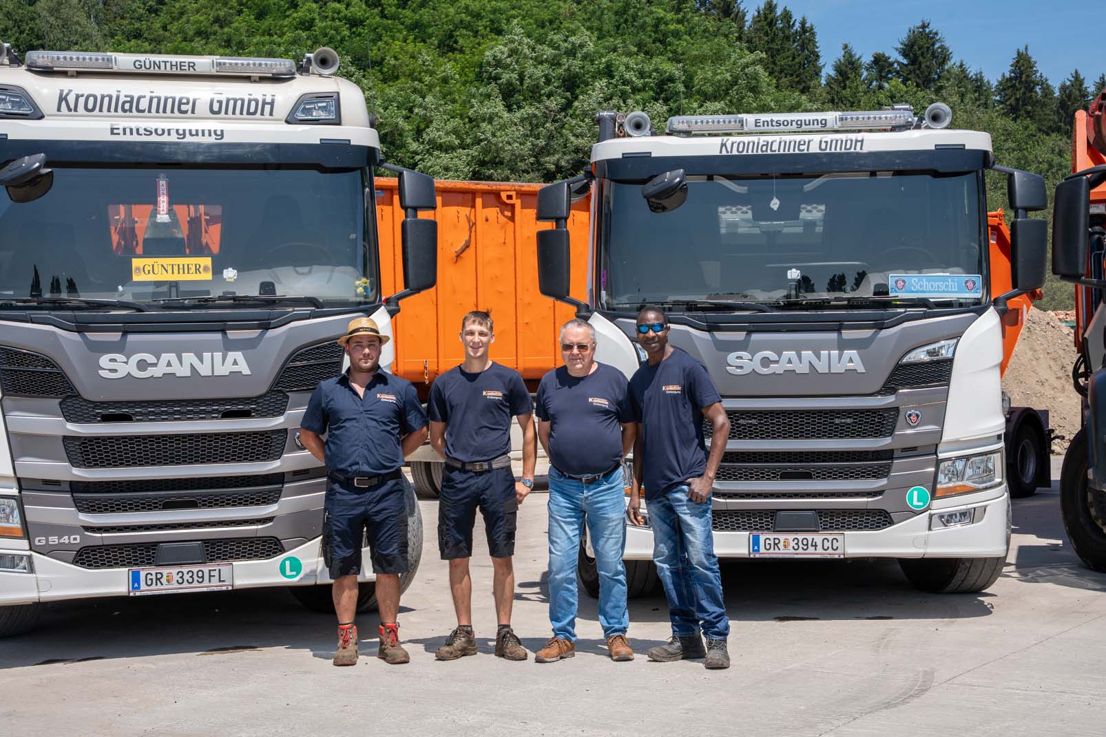 Vier Männer in Arbeitskleidung stehen vor zwei großen weißen Scania LKWs mit orangefarbenen Containern, umgeben von Bäumen.