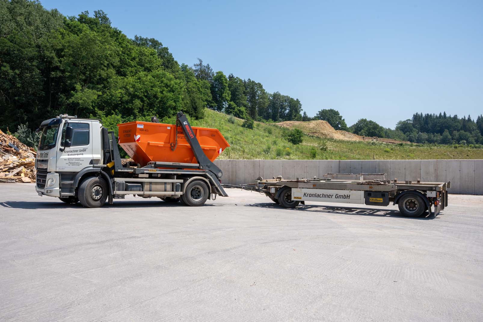 Lastwagen mit orangefarbenem Absetzcontainer und Anhänger auf Betonfläche vor bewaldetem Hang.