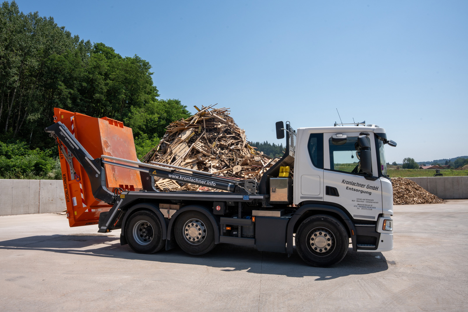 Weißer Entsorgungs-LKW mit orangefarbenem Container vor einem großen Holzabfallhaufen bei klarem Himmel.