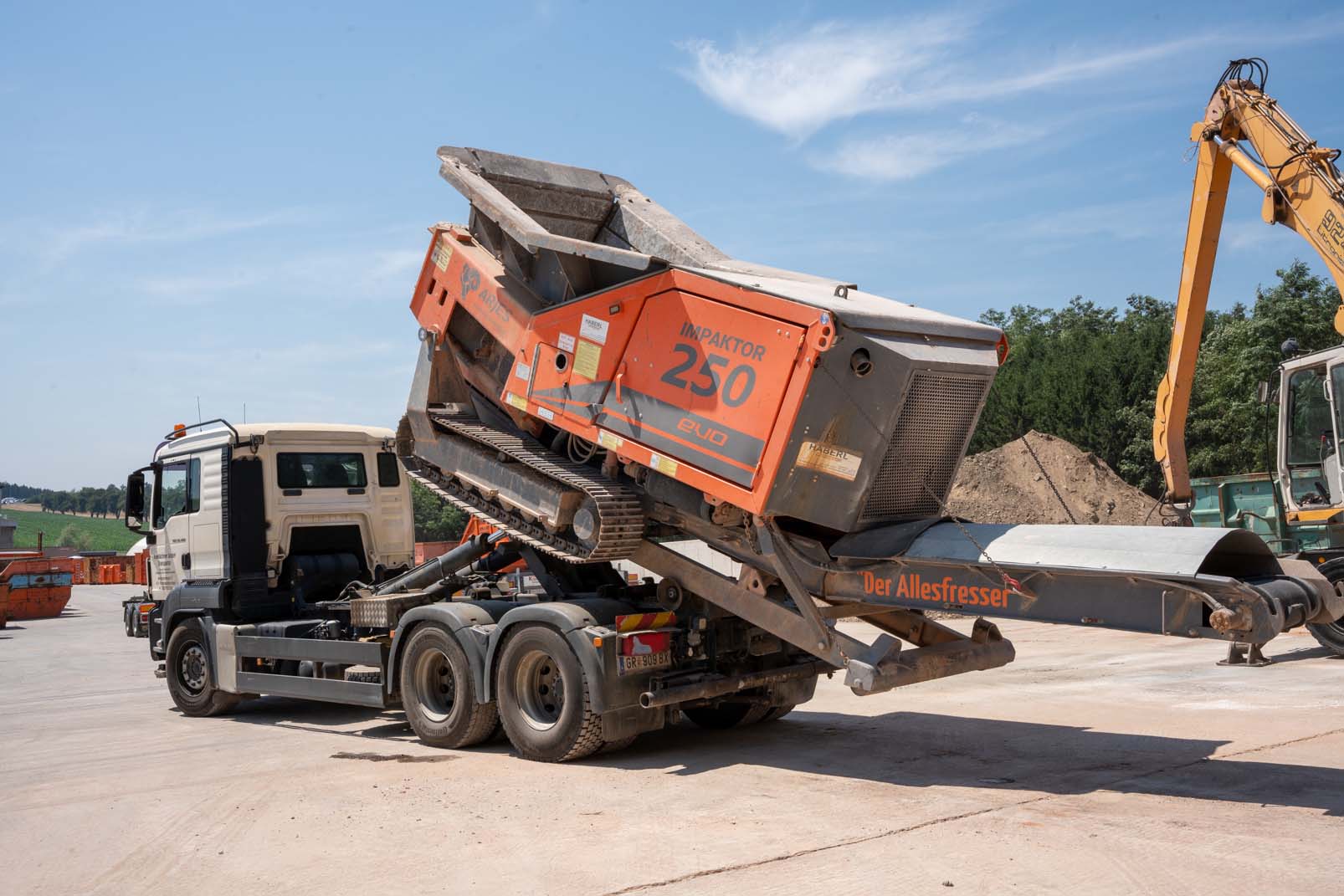 Abfallentsorgungs-LKW mit entladenem orangefarbenem IMPAKTOR 250 evo Gerät auf Baustelle bei klarem Himmel.