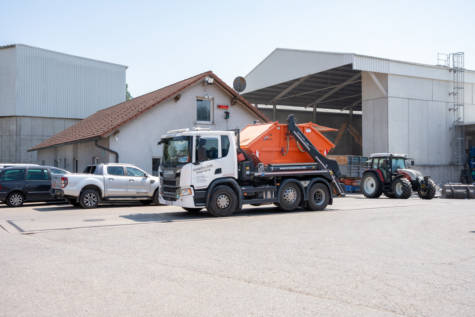 Ein Müllentsorgungs-Lkw der Kronlachner GmbH mit orangefarbener Abfallmulde steht vor einem Industriegebäude neben einem Traktor und mehreren Autos.