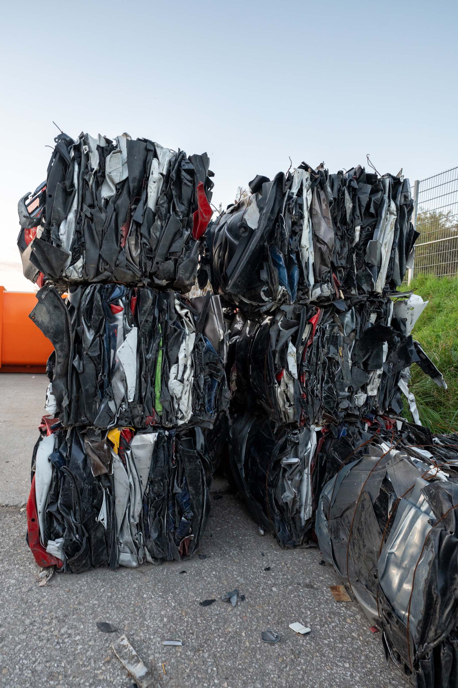 Gestapelte, zusammengedrückte und verdichtete Autowracks auf einem Parkplatz neben einem Zaun und Gras.