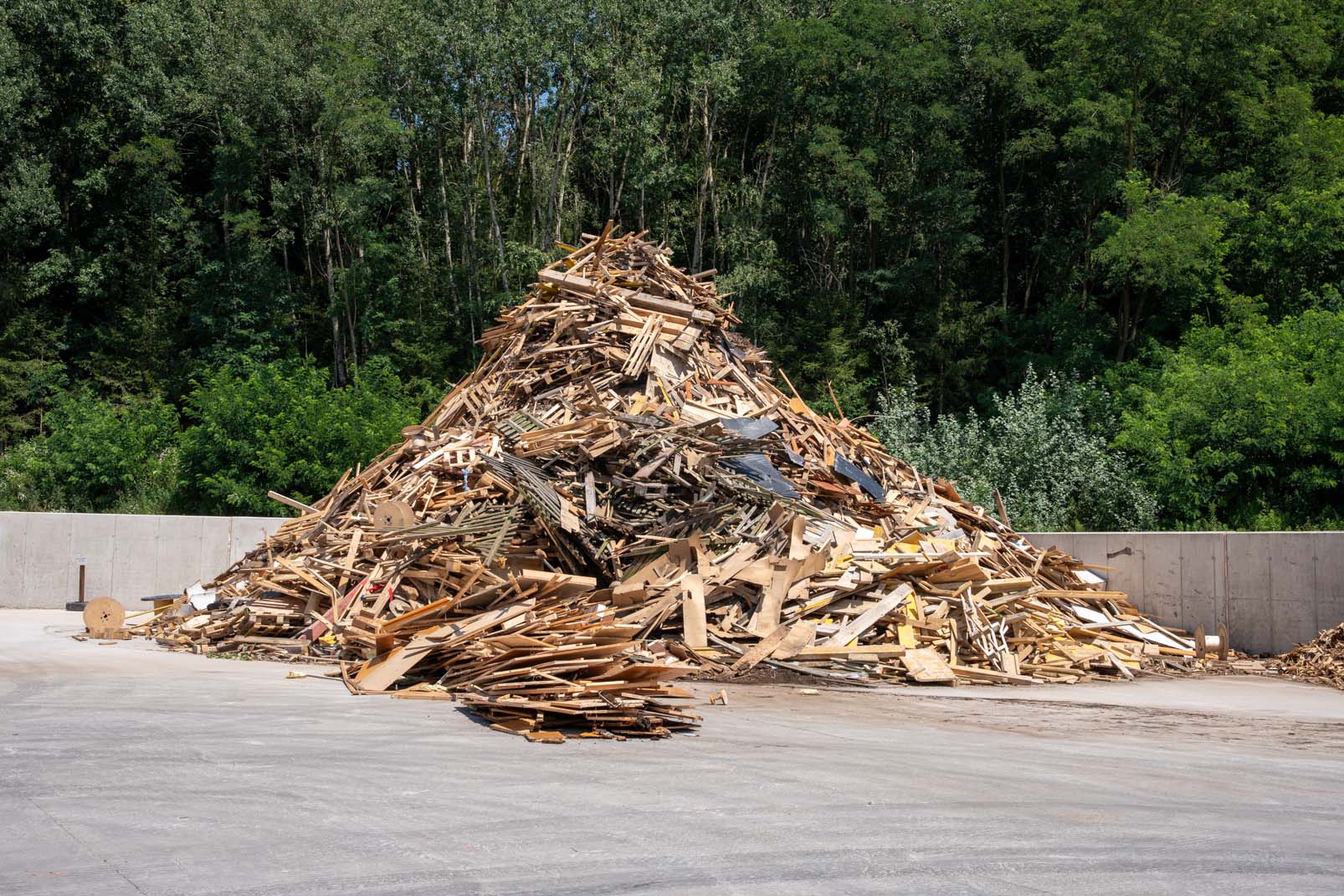 Großer Haufen aus Holzabfällen und Paletten auf Betonboden vor einem Wald.