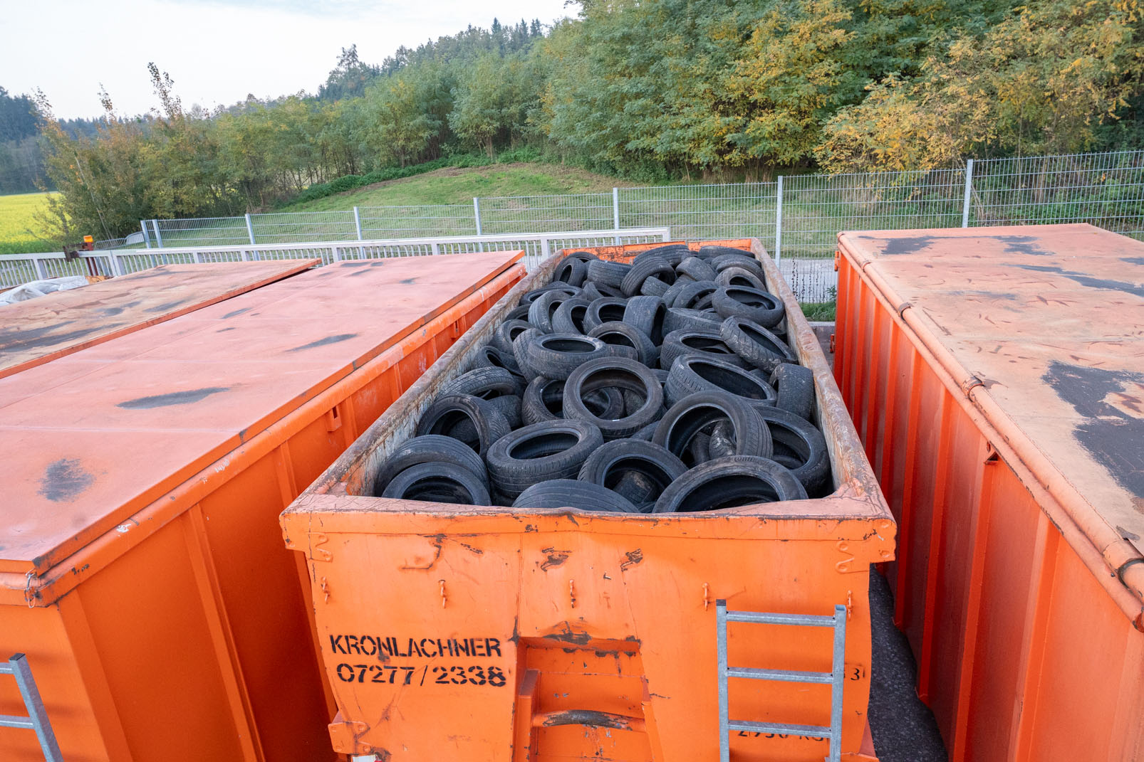 Großer oranger Container gefüllt mit gebrauchten schwarzen Autoreifen, umgeben von weiteren orangen Containern und Bäumen im Hintergrund.