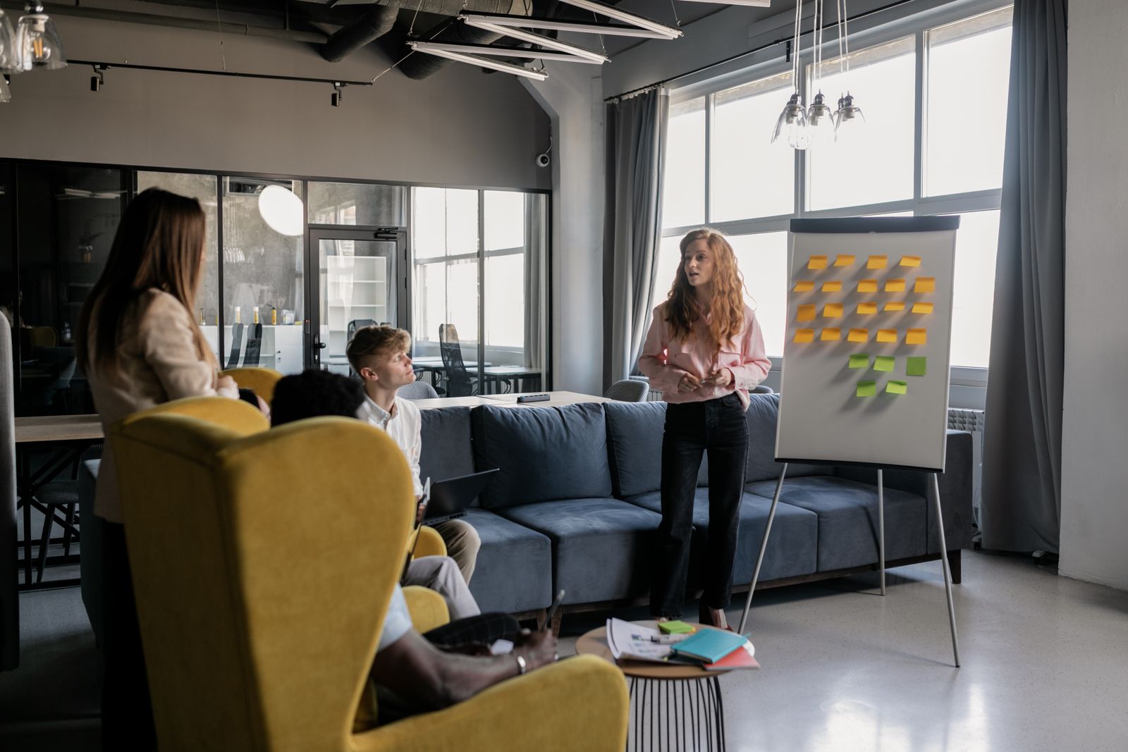 Une femme debout près d'un tableau blanc couvert de post-its échange avec trois collègues lors d'une réunion d'équipe