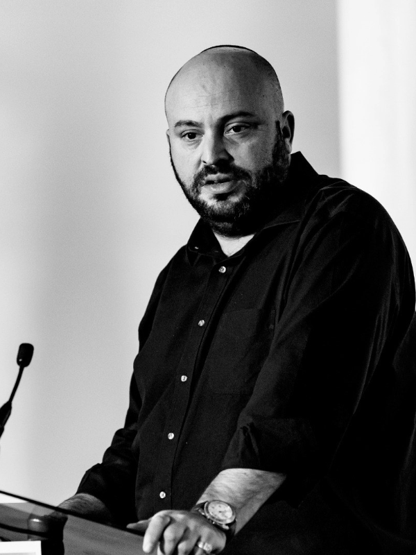 Bearded man in a black shirt speaking at a podium with a microphone.