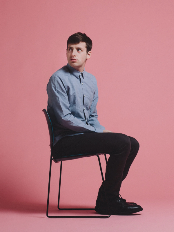 Young man in a blue button-up shirt and black pants sitting on a black chair against a pink background, looking to the side.