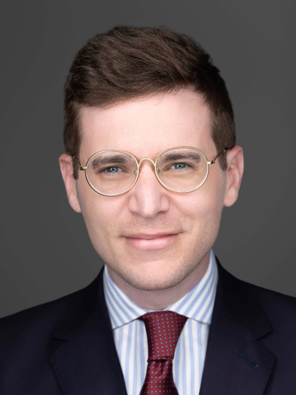 Portrait of a man with short brown hair wearing round gold glasses, a navy blazer, a light blue and white striped shirt, and a red patterned tie.