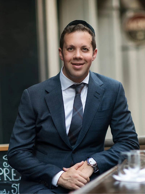 Smiling man dressed in a dark blue suit and tie sitting at a table outdoors.