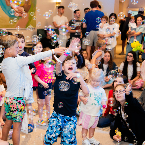 Children playing and reaching for bubbles indoors with adults and other kids watching in the background.