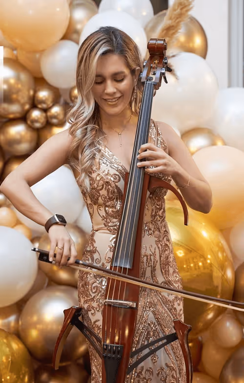 Woman in a sparkling rose gold dress playing an electric cello in front of gold and white balloons.