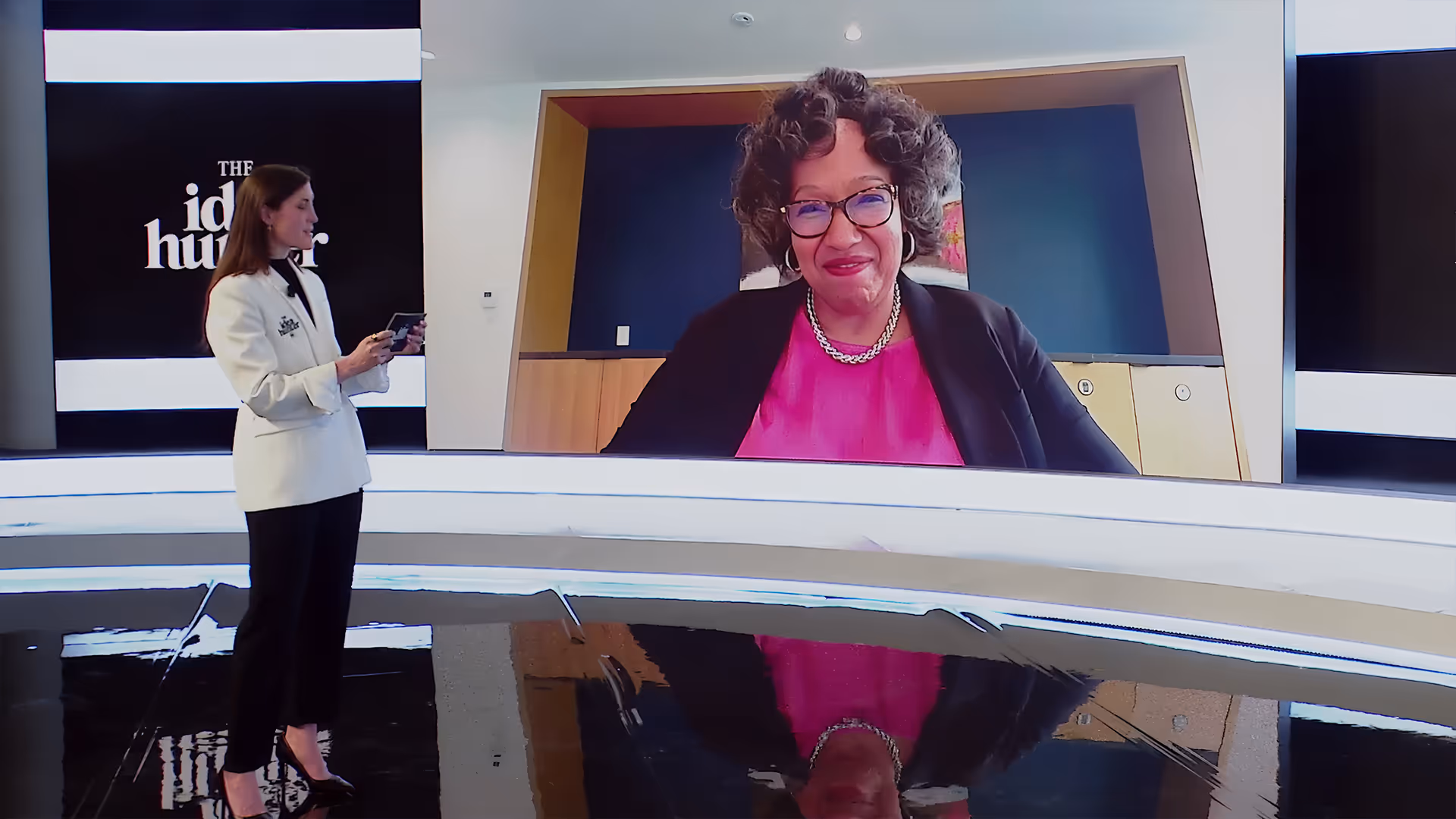 Woman in white blazer standing and holding phone with another woman in a pink top and black blazer smiling on a large screen in a TV studio.