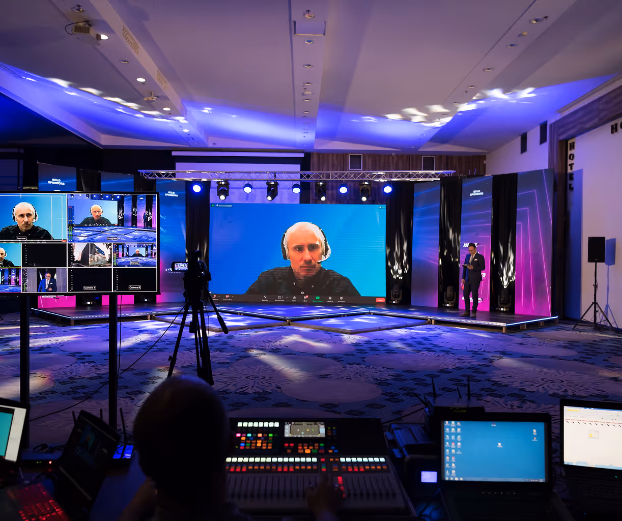 Conference room with a large screen showing a man in a headset on a video call, a smaller monitor displaying multiple video feeds, and a control desk with equipment in the foreground.