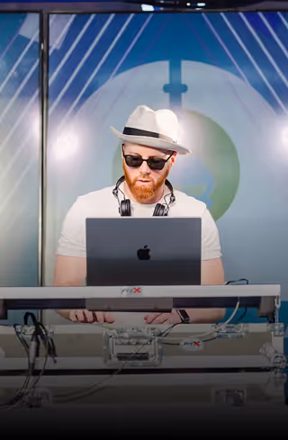 Man with a beard wearing sunglasses and a white hat, using a laptop at a DJ setup.