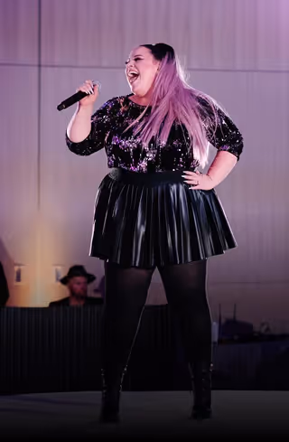 Woman with long pink hair singing on stage holding a microphone, wearing a black sequined top and pleated skirt.