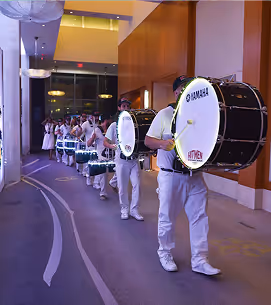 Line of drummers in white uniforms playing bass drums indoors in a hallway.
