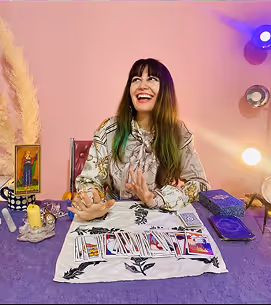 Smiling woman with long hair sitting at a table with tarot cards and candles, appearing to perform a tarot reading.