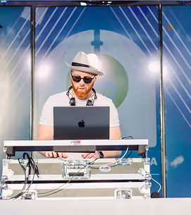 DJ wearing a white hat and sunglasses mixing music on a laptop and keyboard at an outdoor event.