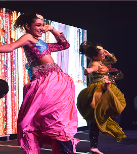 Two female dancers performing on stage wearing colorful traditional costumes, one in pink and the other in yellow.