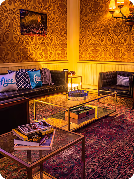 Cozy living room with vintage patterned wallpaper, dark leather sofas, glass coffee tables, and warm lighting.