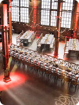 Event space with long banquet tables set with plates, glasses, and floral centerpieces under large industrial windows and red lighting.