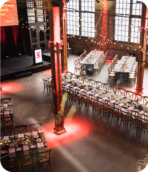 Interior of an event space with long tables set for dining, string lights, large windows, and a stage with a podium.