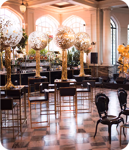 Elegant event space with tall gold and clear balloon decorations, high bar stools, and polished marble floor.