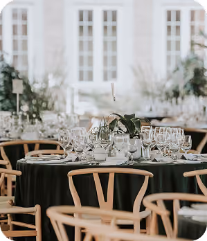 Round table set with glassware and plates, surrounded by wooden chairs at a bright event space.