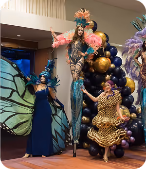 Three performers in elaborate costumes: butterfly, gold balloon dress, and feathered stilt walker.