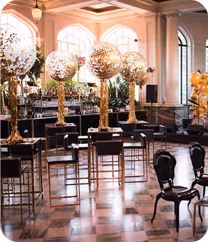 Elegant event space with gold balloon centerpieces on tall tables and black chairs on marble floor.