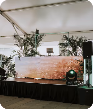 Indoor event stage with wooden podium, green spotlight, and tall potted plants under a white tent ceiling.