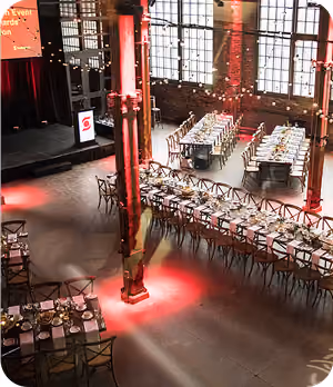 Interior event space with long dining tables set with plates and chairs under large windows.