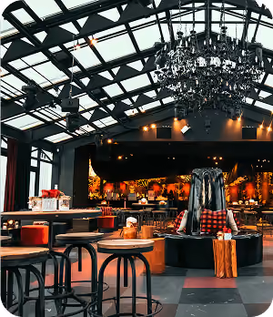 Spacious modern lounge with stools, tables, chandelier, and decorative cushions under a glass ceiling.