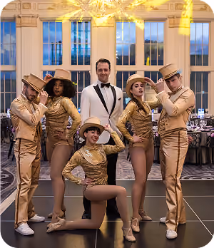 Five performers in gold costumes and hats pose with a man in a white tuxedo in a ballroom.