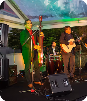 Three musicians performing on stage under a transparent tent at night, playing upright bass, congas, and guitar.