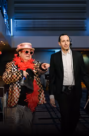 A man in a black suit walking beside a person in shiny silver pants, red feather boa, and pink hat.