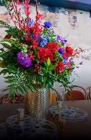 Vibrant bouquet of red, purple, and blue flowers with greenery in a textured gold vase.