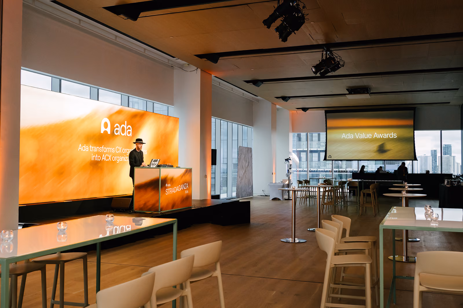 Man in hat standing at a lit podium with orange Ada branding in a modern event space with tables and city views.