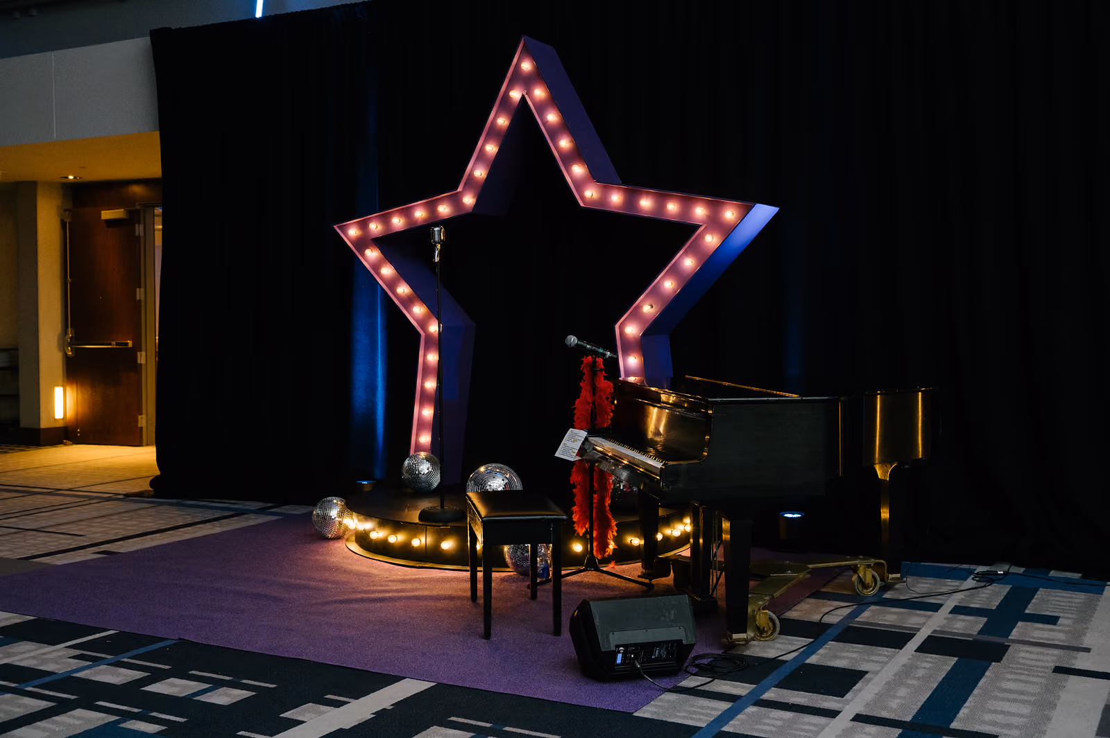 Black grand piano and stool on purple carpet under a lighted star frame with disco balls and microphone.