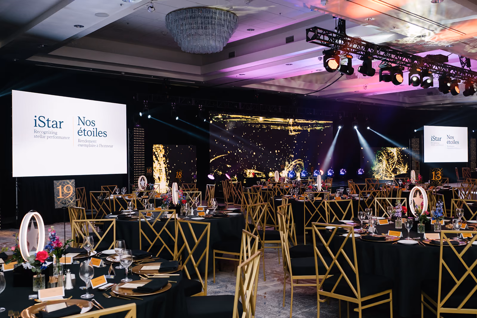 Gala event venue with round tables, gold-trimmed chairs, and illuminated stage and screens.