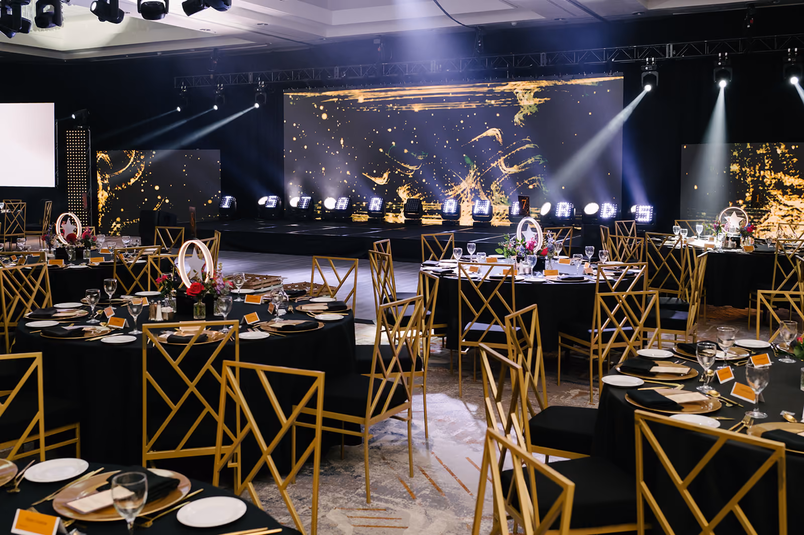 Elegant event space with gold chairs, black-covered round tables, floral centerpieces, and a lit stage.