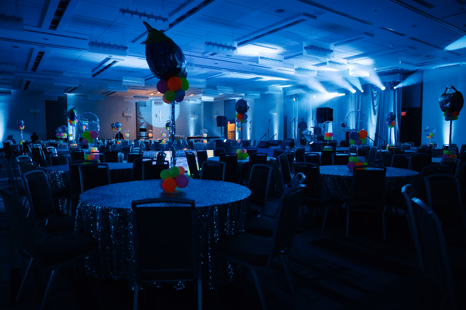 Dark event hall with round tables covered in sparkle cloths and colorful balloon centerpieces, stage lights on.