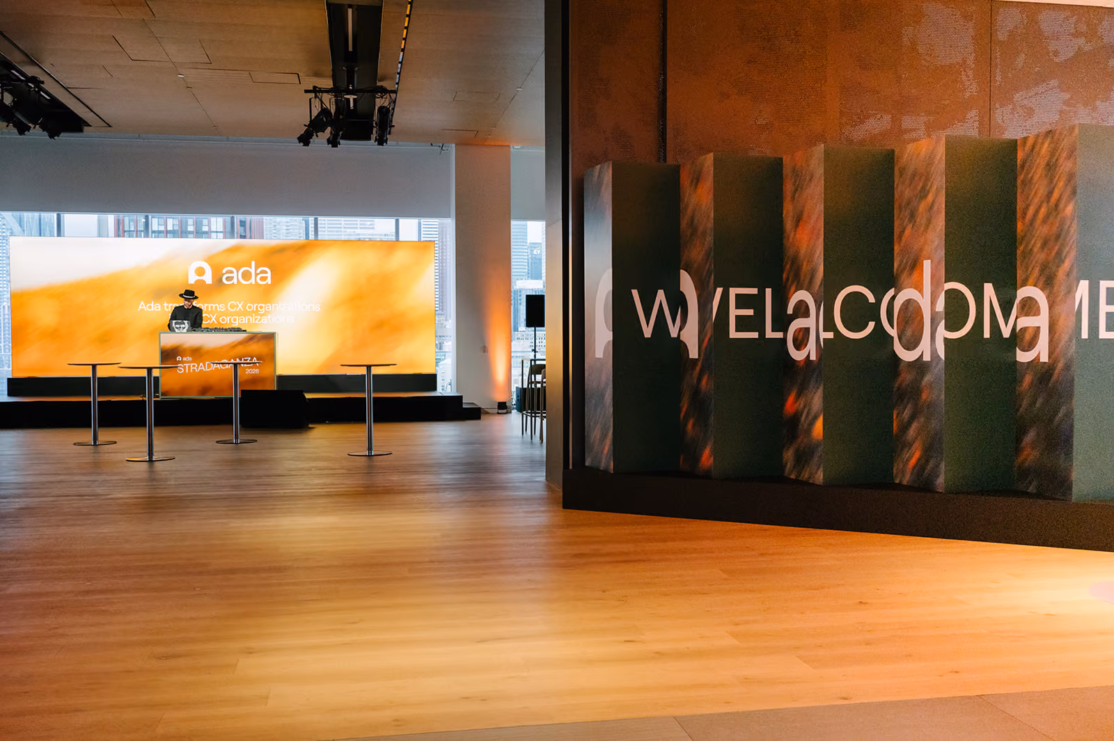 Modern event space with large orange screen displaying Ada logo and a person standing behind a podium.