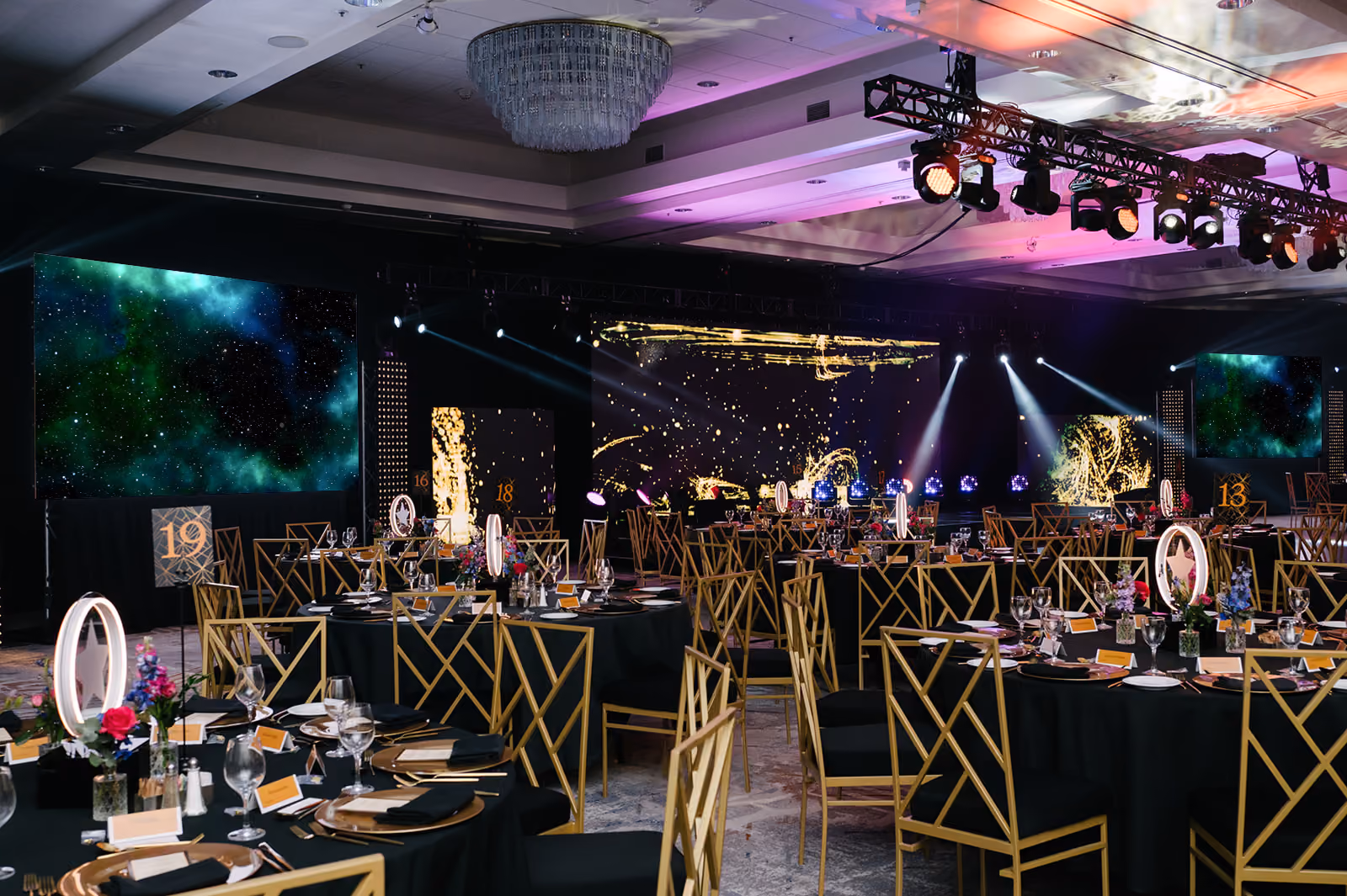 Banquet hall with round tables, gold chairs, black linens, floral centerpieces, and large lit stage screens.