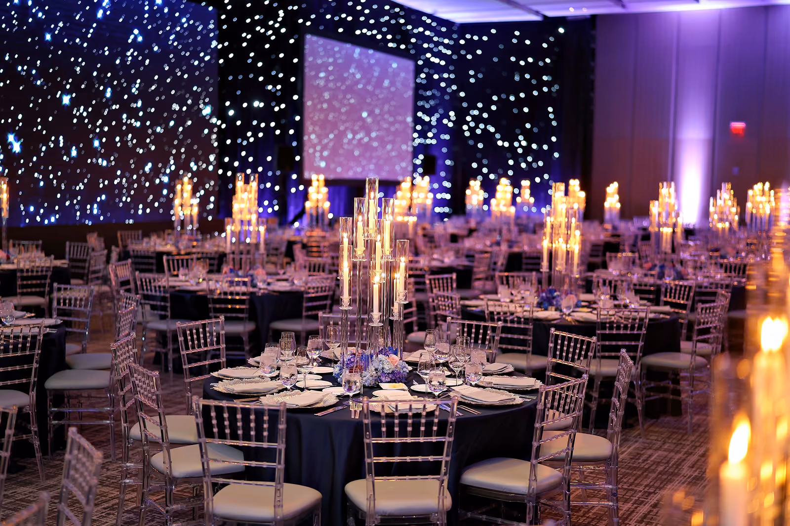 Elegant banquet hall with round tables, glass chairs, candles, and starry light decorations on walls.