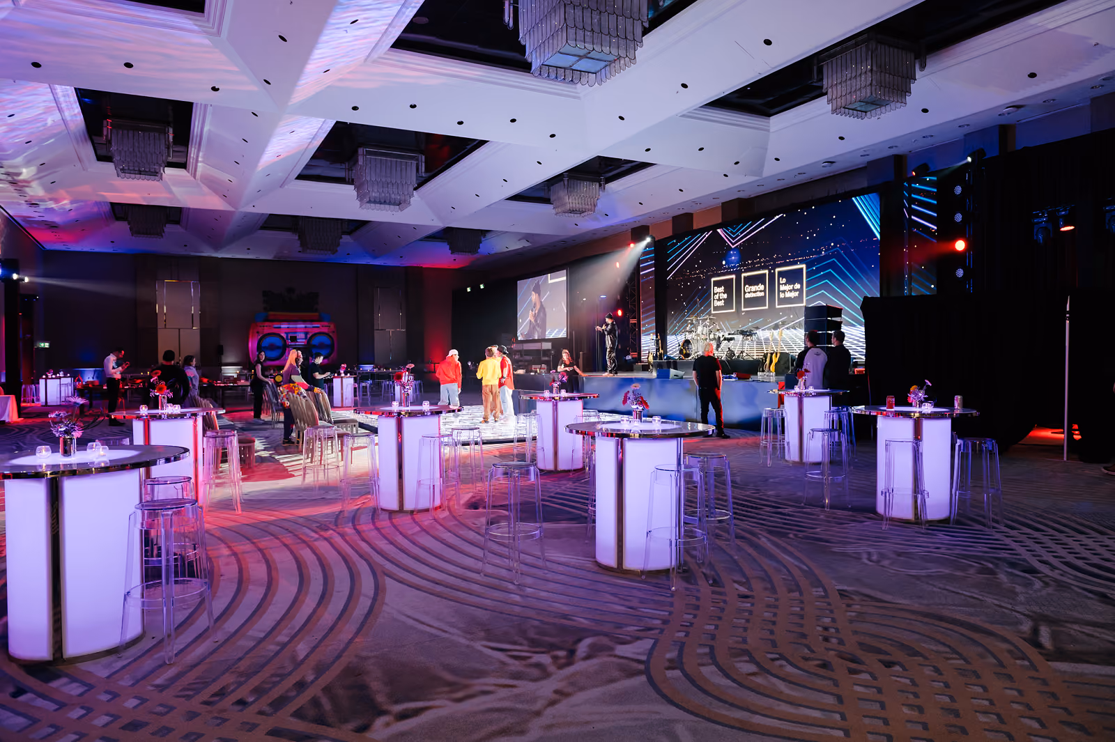 Event hall with illuminated cocktail tables, clear stools, and a stage with lights and people gathering.
